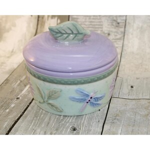 May include: A ceramic jar with a lavender lid and a light green base. The base features embossed leaves, flowers, and a dragonfly. The lid is topped with a leaf-shaped handle. The jar sits on a wooden surface.