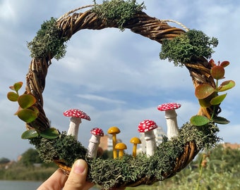以下が含まれることがあります： 編まれた小枝と苔でできたリースで、中央に赤と白のキノコと黄色のキノコの群れがあります。