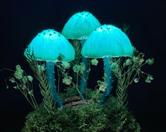 Puede incluir: Tres hongos azules brillantes con puntos blancos están dispuestos en una escena de bosque musgoso. Los hongos están hechos de resina y están iluminados desde adentro, creando un efecto mágico. La escena está colocada sobre una base de madera con musgo y flores secas.