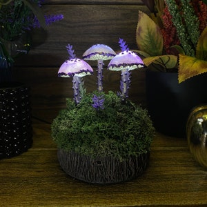 May include: Three purple and white mushroom shaped lamps with glowing lights sit on a bed of green moss. The lamps are arranged in a cluster on a dark brown base.