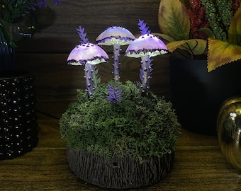 Puede incluir: Tres lámparas con forma de hongo de color morado y blanco con luces brillantes se encuentran sobre un lecho de musgo verde. Las lámparas están dispuestas en un grupo sobre una base marrón oscuro.
