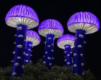 Puede incluir: Un grupo de cinco lámparas con forma de hongo de color morado y blanco con tallos azules y puntos blancos. Las lámparas están dispuestas en semicírculo sobre un lecho de musgo verde.