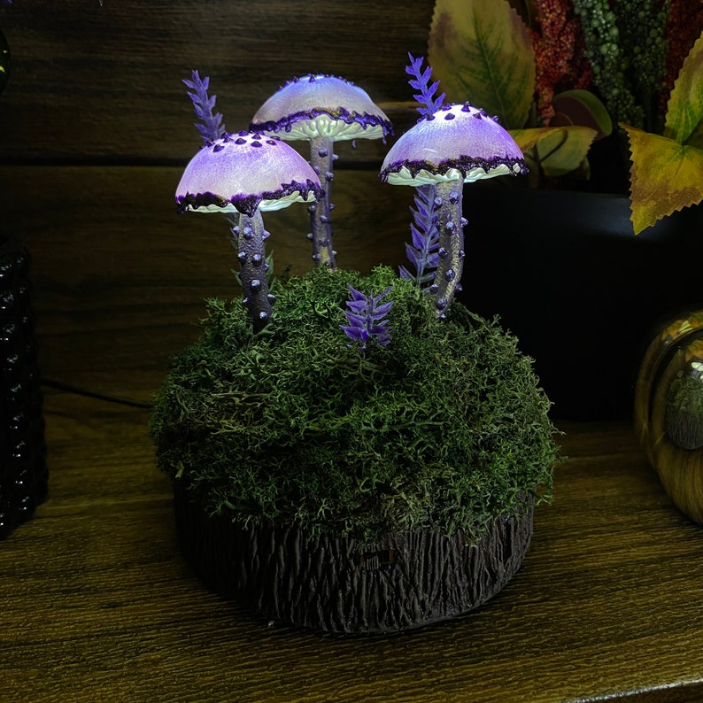May include: A purple and white mushroom lamp with a mossy base. The lamp has three mushrooms with white tops and purple stems. The base is covered in green moss and has a small purple flower.