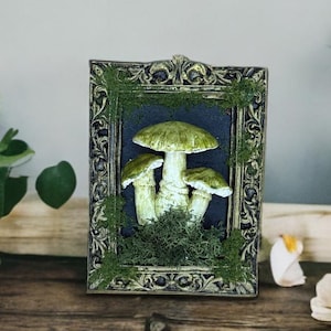 May include: A framed art piece featuring a cluster of green mushrooms. The mushrooms are set against a dark background and surrounded by moss. The ornate frame has gold accents and a dark finish. The artwork is displayed on a wooden surface.