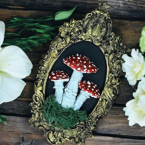 May include: A framed artwork featuring three red and white mushrooms with white stems and green moss on a black background. The frame is gold and ornate.