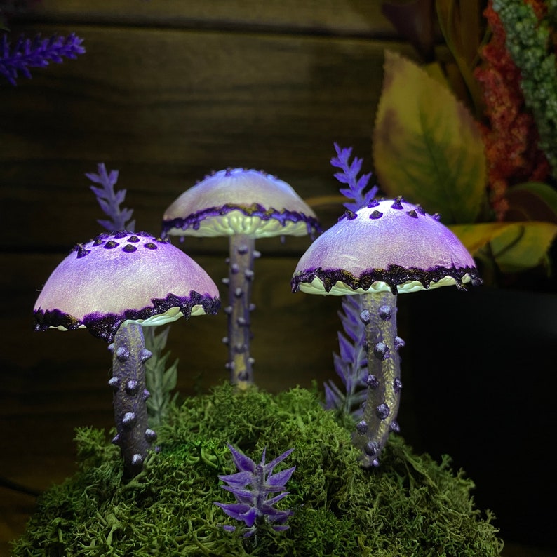 May include: Three purple and white mushroom-shaped lamps with a glittery finish sit on a bed of green moss. The lamps are illuminated with a soft, white light.
