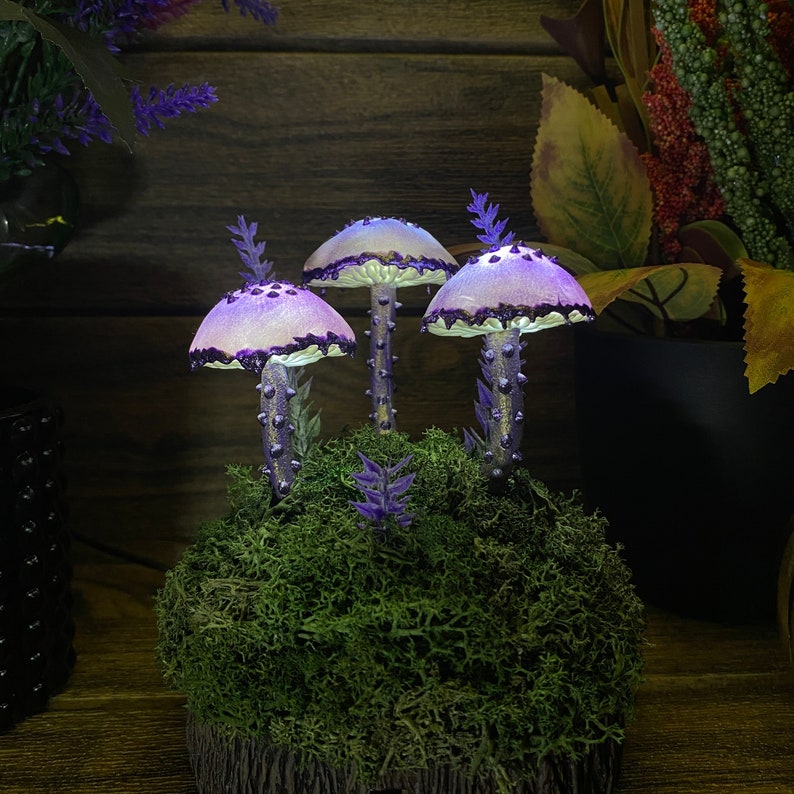May include: Three purple and white mushroom shaped fairy lights with a mossy base. The mushrooms are lit up and have a whimsical feel.