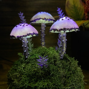 May include: Three purple and white mushrooms with black glitter accents are arranged on a bed of green moss. The mushrooms have a whimsical and magical feel.