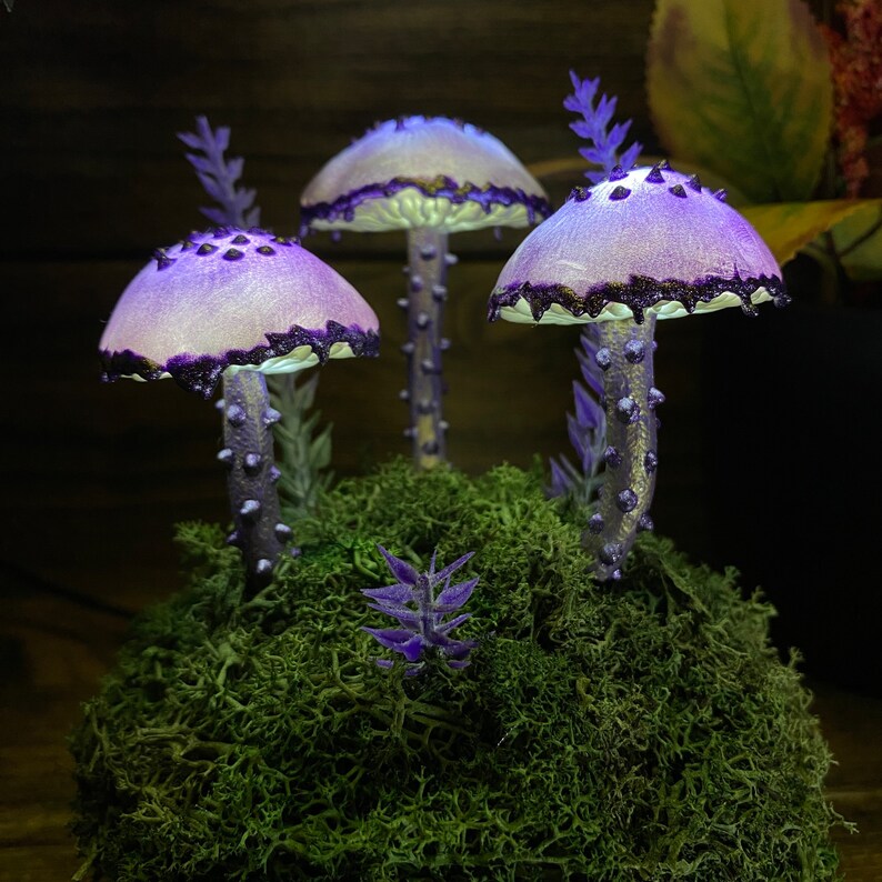 May include: Three purple and white mushrooms with black glitter accents are arranged on a bed of green moss. The mushrooms have a whimsical and magical feel.