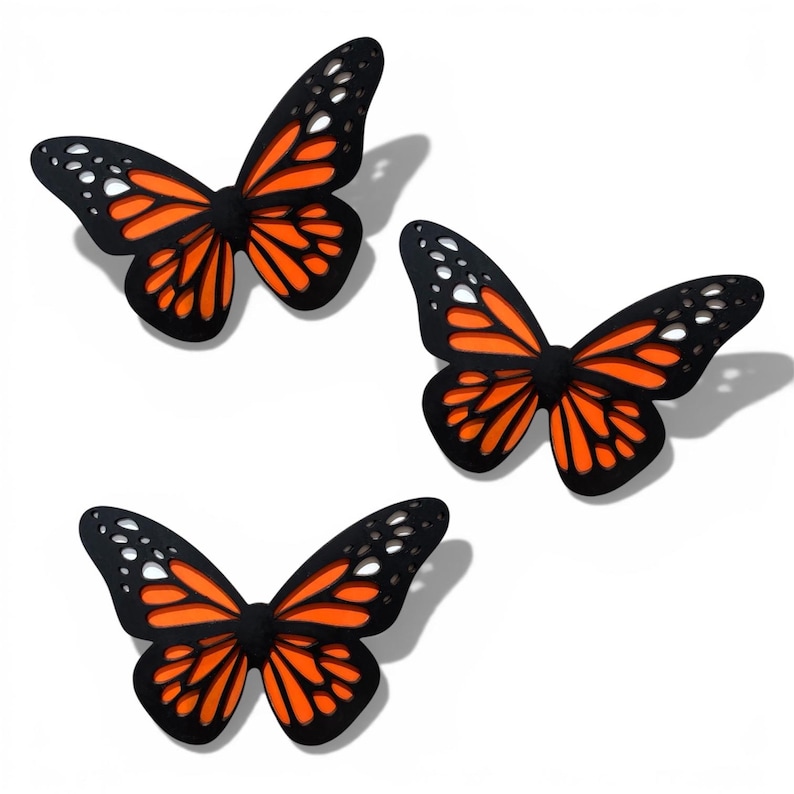 May include: Three decorative butterfly ornaments. Each butterfly features a black outline with orange and white wing details. The butterflies are arranged on a white background, with shadows suggesting they are slightly raised.