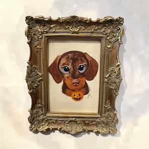 May include: A painting of a brown dachshund with large eyes wearing an orange jack-o'-lantern pendant, set within an ornate, antique-style gold-toned frame with intricate floral and scrollwork detailing along the edges of the rectangular border.