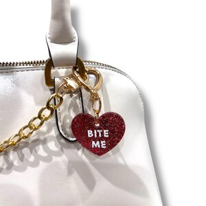 May include: A red glitter heart-shaped charm with the words "BITE ME" in white lettering. The charm is attached to a gold chain and clasp, hanging from a white handbag.