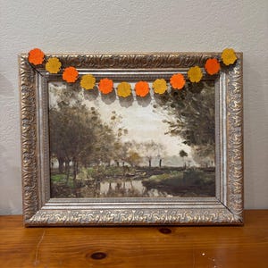 May include: A landscape painting in an ornate, metallic gold-toned frame, featuring a dark, moody scene of trees and a body of water. Draped across the top of the frame is a garland of small, circular felt flowers in alternating shades of orange and yellow.