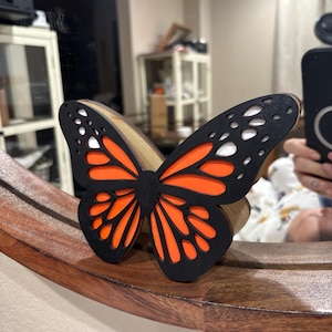May include: A wooden butterfly decoration with black, orange, and white accents. The butterfly is perched on a wooden surface. The butterfly's wings have a detailed cutout design.