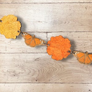 May include: A wooden garland featuring orange butterflies and marigold-like flowers. The butterflies have detailed wing patterns, and the flowers have intricate petal designs. The garland is connected with small metal rings, creating a decorative chain.