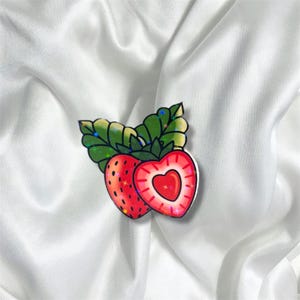 May include: A red and green sticker with a cartoon image of a strawberry with a heart-shaped center. The strawberry is surrounded by green leaves.