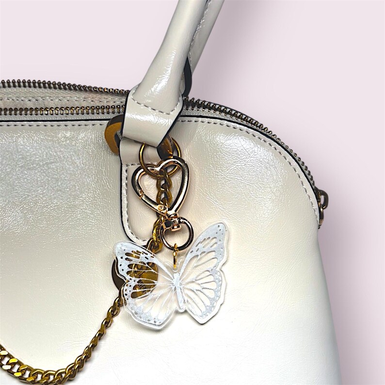 May include: A white handbag with a gold zipper and handle. A gold-colored heart-shaped clasp holds a butterfly charm. The butterfly charm is white with gold accents and hangs from a gold chain.