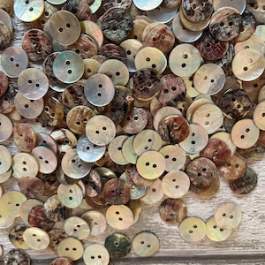 May include: A pile of round, two-hole shell buttons in various shades of brown, beige, and white. The buttons have a natural, iridescent sheen.