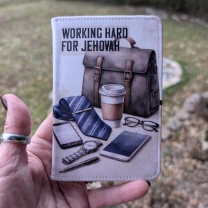 Puede incluir: Una cartera blanca con el texto "WORKING HARD FOR JEHOVAH" y una ilustraci&oacute;n de un malet&iacute;n, corbata, taza de caf&eacute;, reloj, gafas y tel&eacute;fonos inteligentes. La cartera se sostiene en una mano.