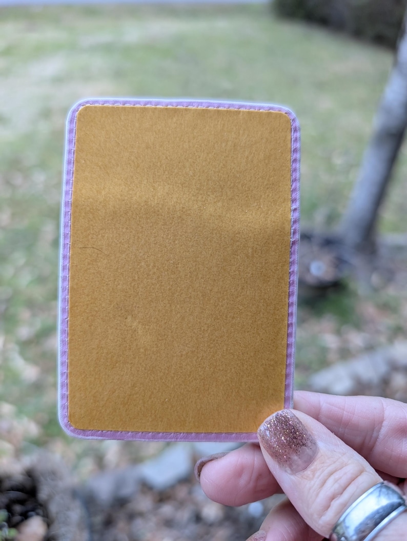 May include: A gold-colored sticker with a pink border. The sticker has a stitched edge.