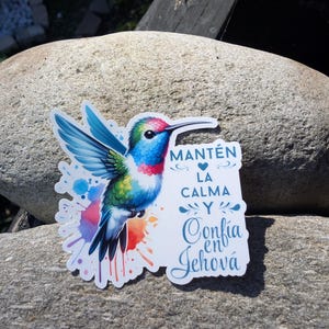 May include: A colorful sticker featuring a hummingbird with blue, green, red, and yellow feathers. The sticker has the text "MANTÉN LA CALMA Y Confía en Jehová" in blue script. The sticker is placed on a gray rock.