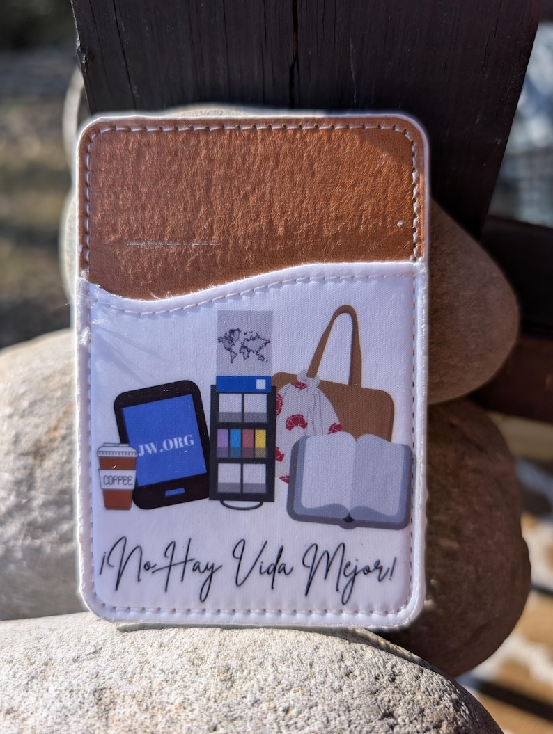 May include: A white phone case with a brown leather flap. The case features a design of a coffee cup, a tablet with the text "JW.ORG", a book, a tote bag, and a magazine. The text "No Hay Vida Mejor!" is printed on the case.