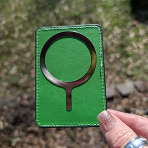 May include: A green leather card holder with a metal ring and a metal stick extending from the ring. The card holder has a stitched border.