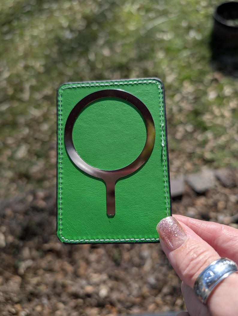 May include: A green leather card holder with a metal ring and a metal stick extending from the ring. The card holder has a stitched border.