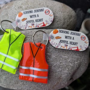 May include: Keychains featuring a neon green and orange vest design, alongside two keychains with the text "SERVING JEHOVAH WITH A JOYFUL HEART". The keychains are set against a gray stone background.