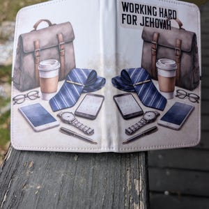 Puede incluir: Una cartera blanca plegable con un dise&ntilde;o impreso que presenta maletines, tazas de caf&eacute;, corbatas, gafas, relojes y tel&eacute;fonos inteligentes. El texto "WORKING HARD FOR JEHOVAH" est&aacute; impreso en la parte superior.