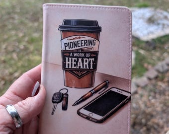 Puede incluir: Un portapasaportes de color rosa claro con una ilustración gráfica de un vaso de café con el texto PIONEERING IS A WORK OF HEART, un juego de llaves de coche, un bolígrafo y un teléfono inteligente. El diseño está impreso en una funda lisa.