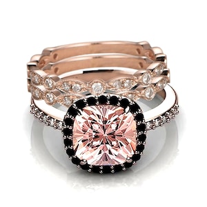 Puede incluir: Un juego de anillos de compromiso de tres piezas en oro rosa con una gran piedra preciosa rosa de talla cojín rodeada por un halo de diamantes negros. La banda está acentuada con diamantes más pequeños.