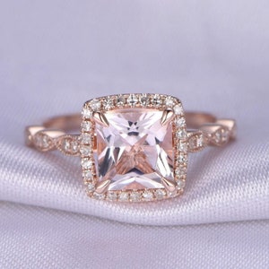 May include: A rose gold engagement ring with a square-shaped morganite center stone surrounded by a halo of smaller diamonds. The band is also adorned with smaller diamonds.