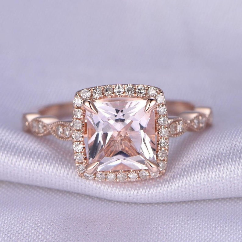 May include: A rose gold engagement ring with a square-shaped morganite center stone surrounded by a halo of smaller diamonds. The band is also adorned with smaller diamonds.