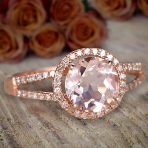 Peut inclure: Bague en or rose ornée d'une grosse pierre rose pâle ronde, entourée d'un halo de petites pierres claires. Le corps de la bague se divise en deux parties, également serties de petites pierres claires. Fond flou.