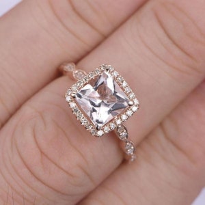 May include: A rose gold engagement ring with a square-shaped center stone surrounded by a halo of smaller diamonds. The band is adorned with a delicate, twisted design.