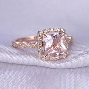May include: A rose gold engagement ring with a square-shaped morganite center stone surrounded by a halo of smaller diamonds. The band is adorned with a delicate, twisted design.