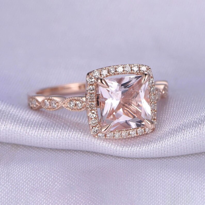 May include: A rose gold engagement ring with a square-shaped morganite center stone surrounded by a halo of smaller diamonds. The band is adorned with a delicate, twisted design.