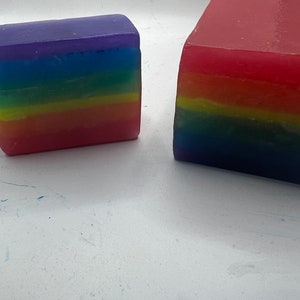 May include: Two colorful, layered soap bars. One is a smaller rectangular shape, and the other is a larger, more square shape. The colors of the soap bars are in a rainbow pattern, with layers of red, orange, yellow, green, blue, and purple.
