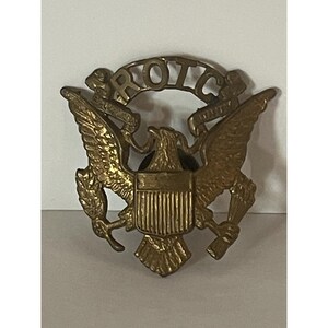 May include: A brass-colored ROTC insignia featuring an eagle with outstretched wings, a shield, and the letters "ROTC" across the top. The eagle holds a bundle of arrows and an olive branch.