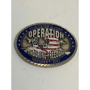 May include: Oval-shaped commemorative coin with a silver border and a blue outer ring with stars. The coin features the text "OUR CAUSE IS JUST" and "OPERATION ENDURING FREEDOM OCTOBER 7, 2001". The center design includes an American flag.