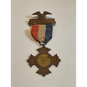 Puede incluir: Medalla de recuerdo de bronce con un águila y la palabra "SOUVENIR" sobre una cinta tricolor. La medalla en forma de cruz tiene un emblema central dorado y el año "1898" inscrito en la parte inferior.