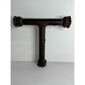Puede incluir: Una herramienta vintage en forma de T de metal marrón oscuro. La herramienta tiene una barra horizontal con extremos hexagonales y una barra vertical que se extiende hacia abajo. La palabra "SOLAT" está estampada en la barra horizontal. La herramienta parece desgastada.