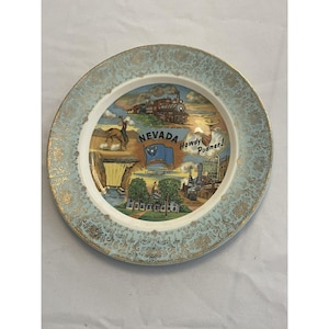 May include: A decorative plate with a light blue rim and gold filigree design. The center features a colorful illustration of Nevada landmarks, including a train, cowboy, and the state flag. Text includes "Nevada" and "Howdy Podner!"