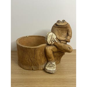 May include: A tan-colored planter shaped like a tree stump with a frog figure sitting on the side. The frog has a textured brown body and is wearing white sneakers. The interior of the stump is hollow and circular, providing space for plants.
