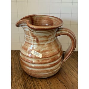 Puede incluir: Jarra de cerámica hecha a mano con asa y boquilla. La jarra es de color marrón claro con rayas horizontales marrones oscuras. Una etiqueta en la parte delantera dice "Annapolis Pottery 40 State Circle."