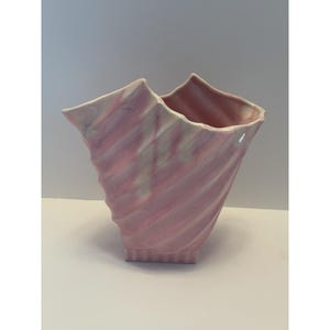 May include: A pink ceramic vase with a unique, twisted design. The vase has a flared top and a textured, ridged surface. The interior is a darker shade of pink. The vase is a decorative item.