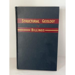 May include: A hardcover book titled "STRUCTURAL GEOLOGY by BILLINGS" in gold lettering on a dark blue cover. The title is centered on a red band with gold trim. The book has a dark blue spine.
