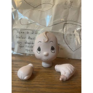 May include: A ceramic baby doll head with a swirl of hair, and two detached hands. The head has large eyes and a serene expression. A pattern for the doll is visible in the background. The text on the pattern says "Make a 5 cm Center Back".