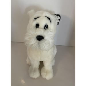 Peut inclure: Une peluche blanche en forme de chien avec des yeux, un nez et des sourcils noirs. Le chien a une barbe blanche duveteuse et des détails noirs sur une oreille. Le chien est assis droit, les pattes avant visibles.